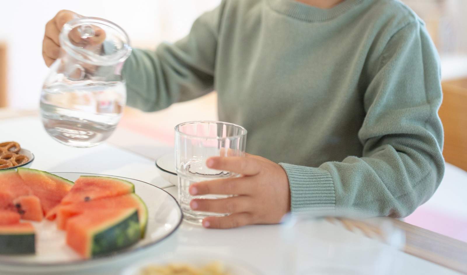 Kind schenkt sich ein Glas Wasser ein. Vor dem Kind liegt ein Teller mit Wassermelonen Stückchen.