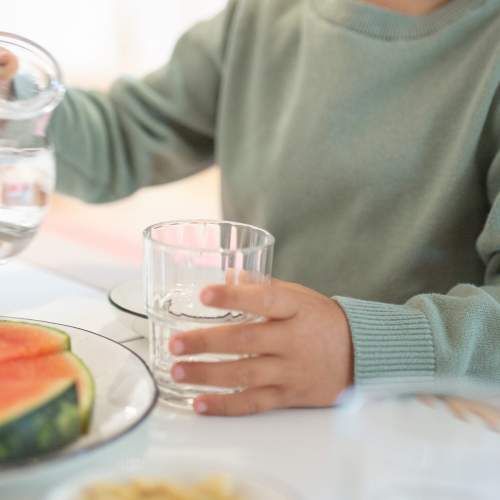 Kind schenkt sich ein Glas Wasser ein. Vor dem Kind liegt ein Teller mit Wassermelonen Stückchen.